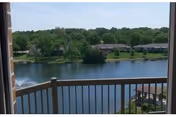 View from a balcony overlooking a lake with a fountain, gazebo, and tree-lined shoreline with low-rise buildings.