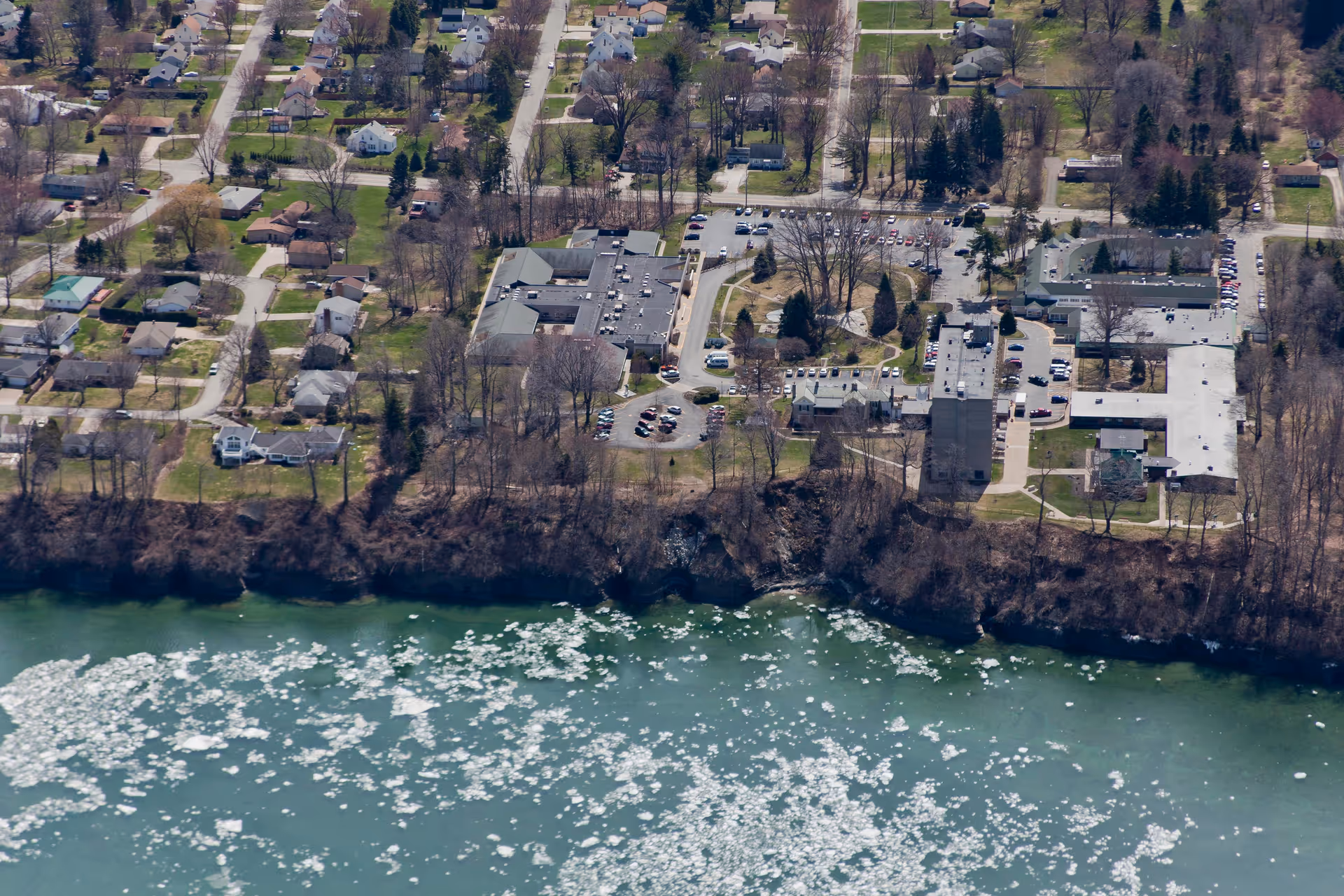 Aerial view of Brevillier Village facility and surrounding neighborhood with houses, trees, parking lots, and a partially frozen body of water in the foreground.