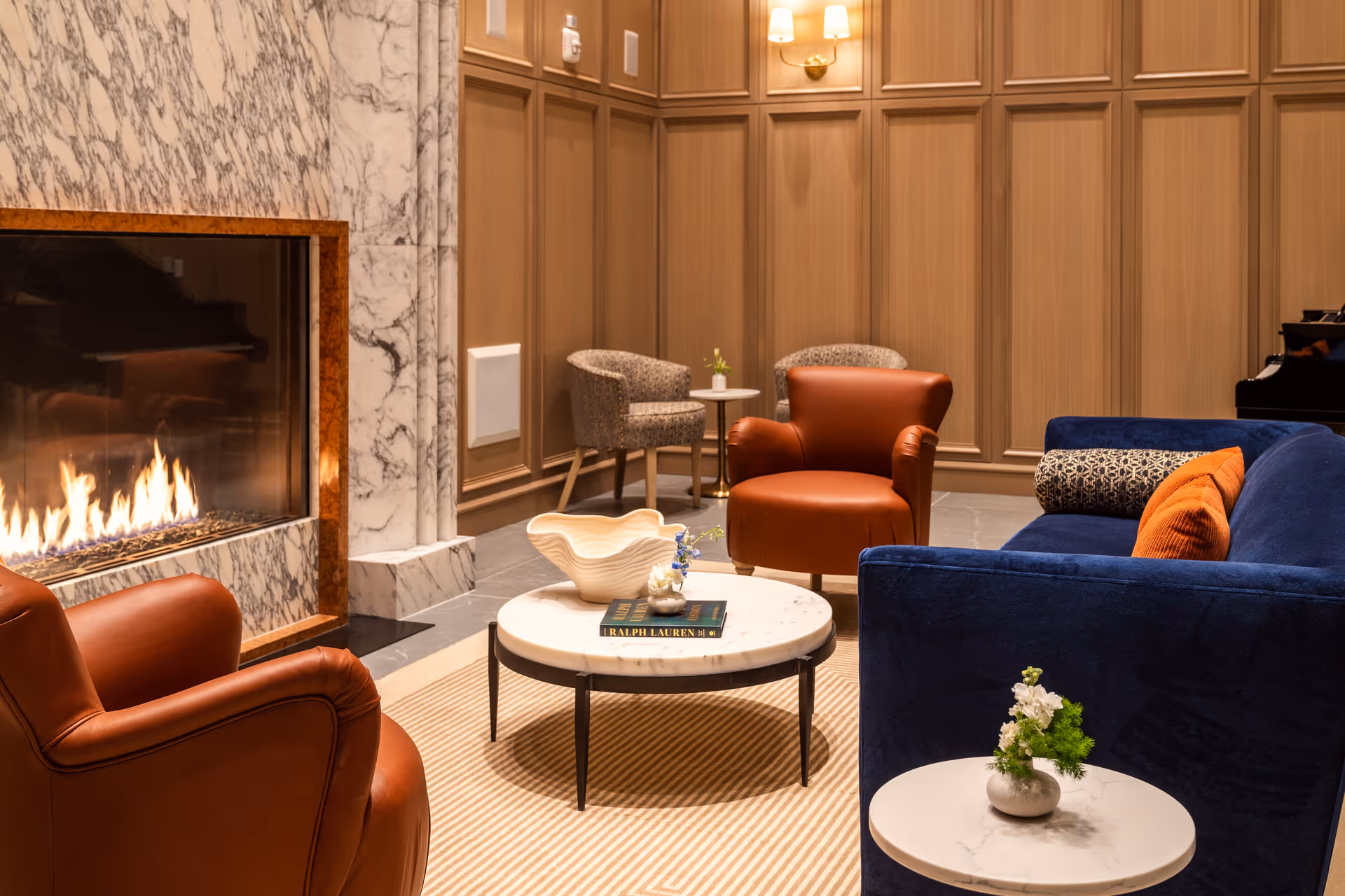 A cozy living room area with a lit marble fireplace on the left, two brown leather armchairs, a blue sofa with orange and patterned cushions, and a round white marble coffee table with decorative items. The walls are paneled in wood, and there are two small round side tables with small flower arrangements. A piano is partially visible in the background.