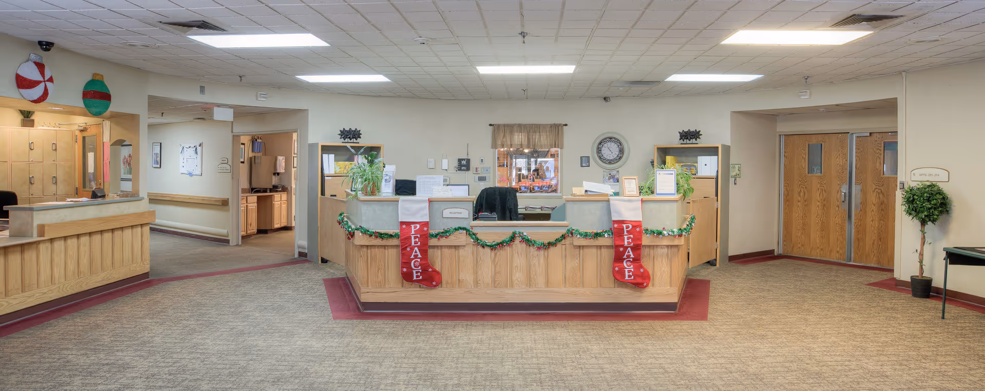 Reception area of a senior living facility decorated for the holidays with two red stockings labeled 'PEACE' hanging on the front desk, garland, and festive ornaments on the wall. The space has beige walls, a carpeted floor, wooden doors, and a clock on the wall behind the desk.