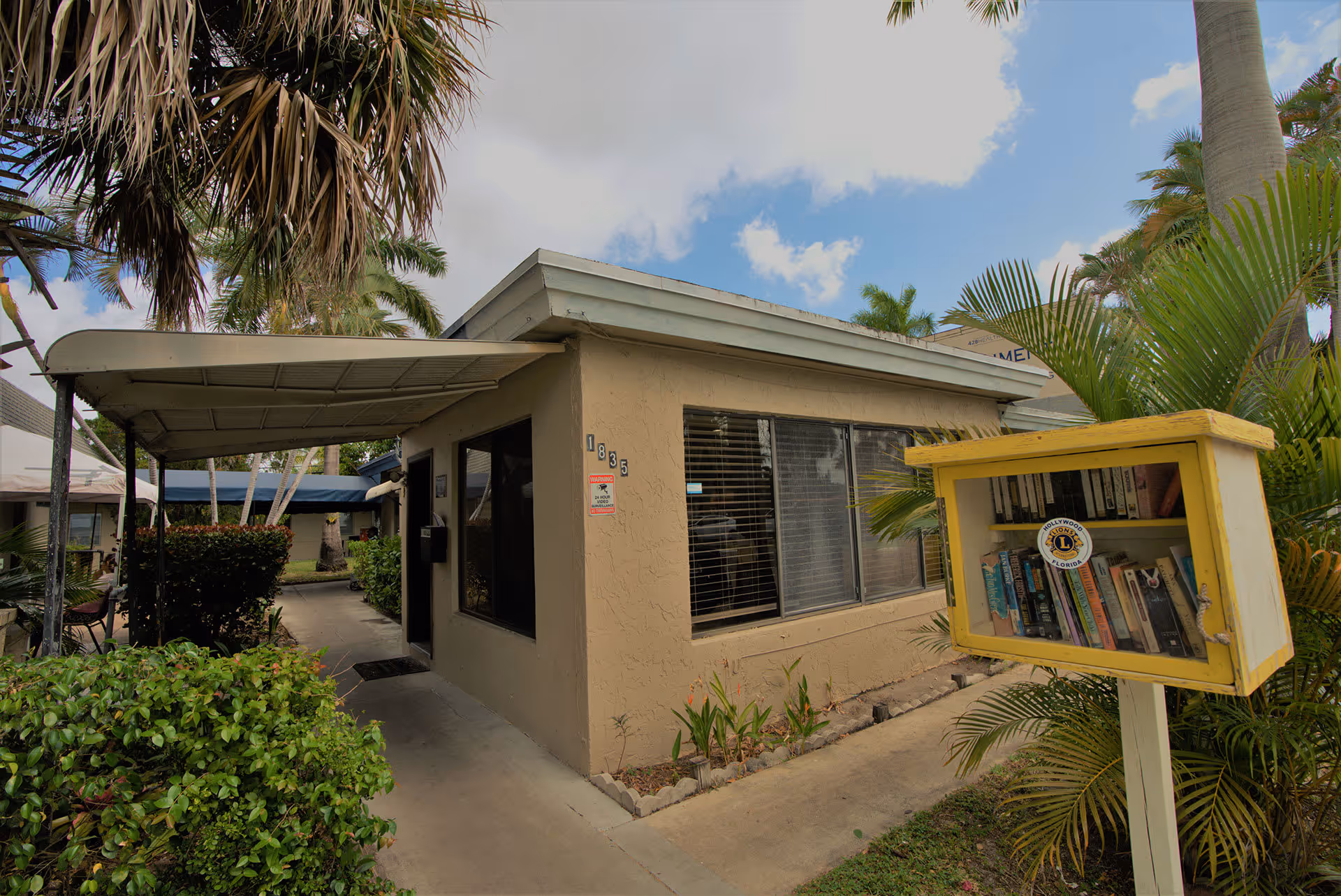 Exterior view of a single-story building with beige walls and large windows, surrounded by palm trees and greenery. A covered walkway leads to the entrance, and a small yellow book exchange box is visible in the foreground with books inside. The sky is partly cloudy.