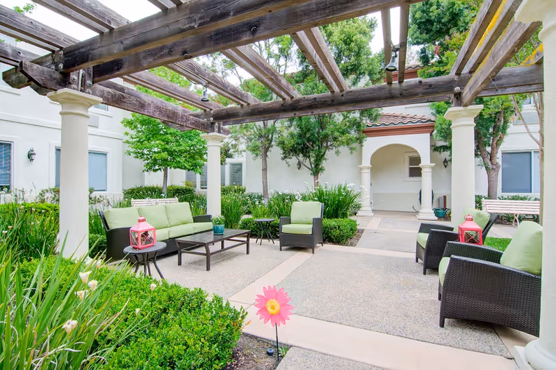 Outdoor seating area with green cushioned chairs and sofas under a wooden pergola surrounded by greenery and trees, located in a courtyard of a building with white walls and arched doorways.