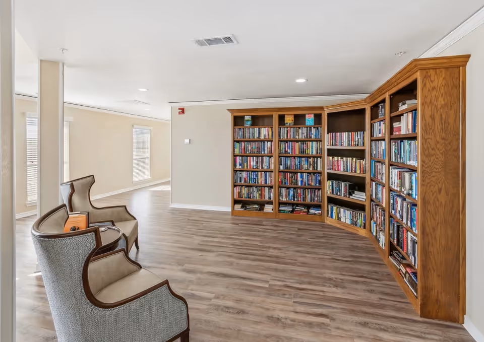 A bright and spacious room with light-colored walls and wood flooring featuring a large wooden bookshelf filled with books along one wall. In the foreground, there are two upholstered armchairs with wooden frames positioned near a small round table. The room has several windows allowing natural light to enter.
