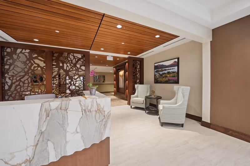 A modern senior living reception lobby with a marble front desk, decorative wood partition and ceiling, and two upholstered chairs with a small table.
