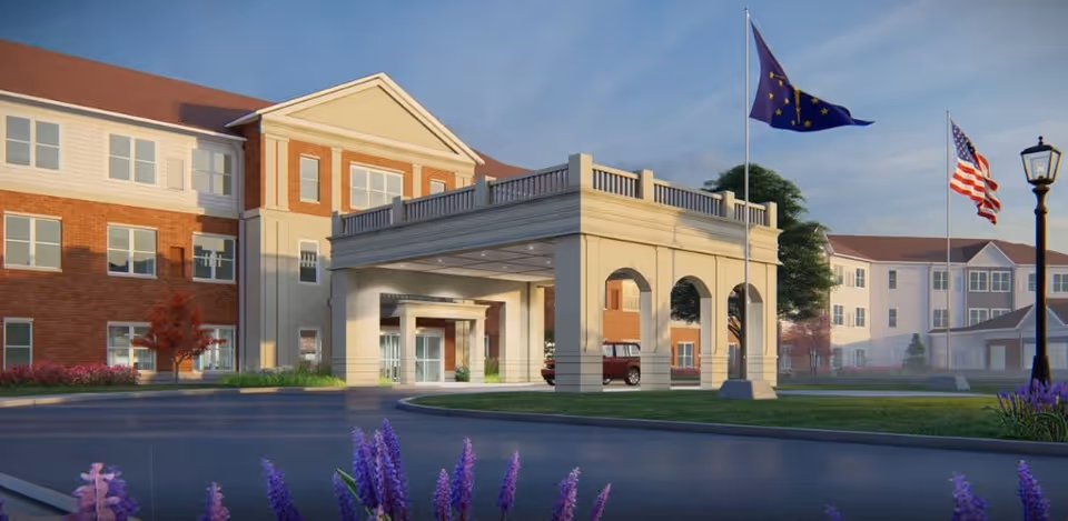 Exterior view of a senior living facility building with a covered entrance, two flagpoles displaying the Indiana state flag and the American flag, a street lamp, and purple flowers in the foreground under a clear sky.