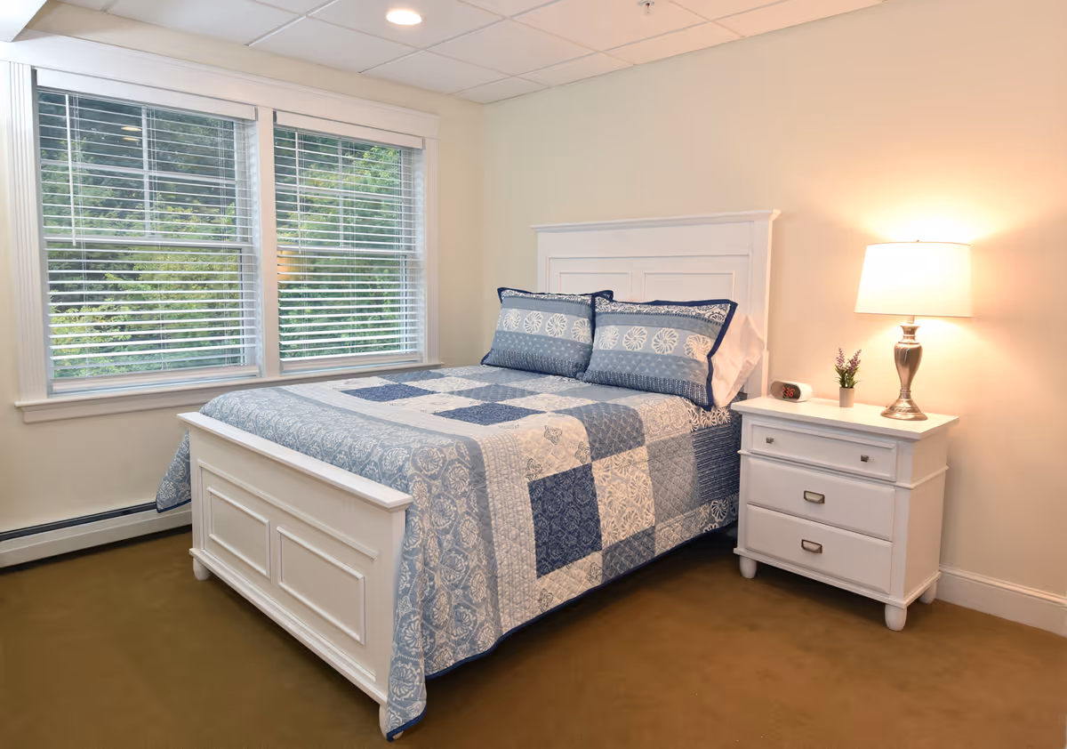 Sunlit bedroom with a white bed dressed in a blue patchwork quilt, a white nightstand with a lamp, and a window with blinds.