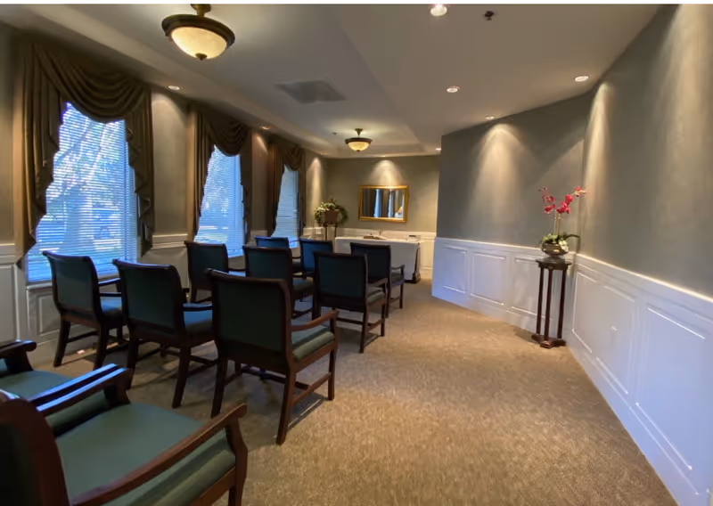Small elegant meeting room with rows of green-upholstered chairs facing a table and mirror, windows with draped valances along one wall, and soft wall lighting.