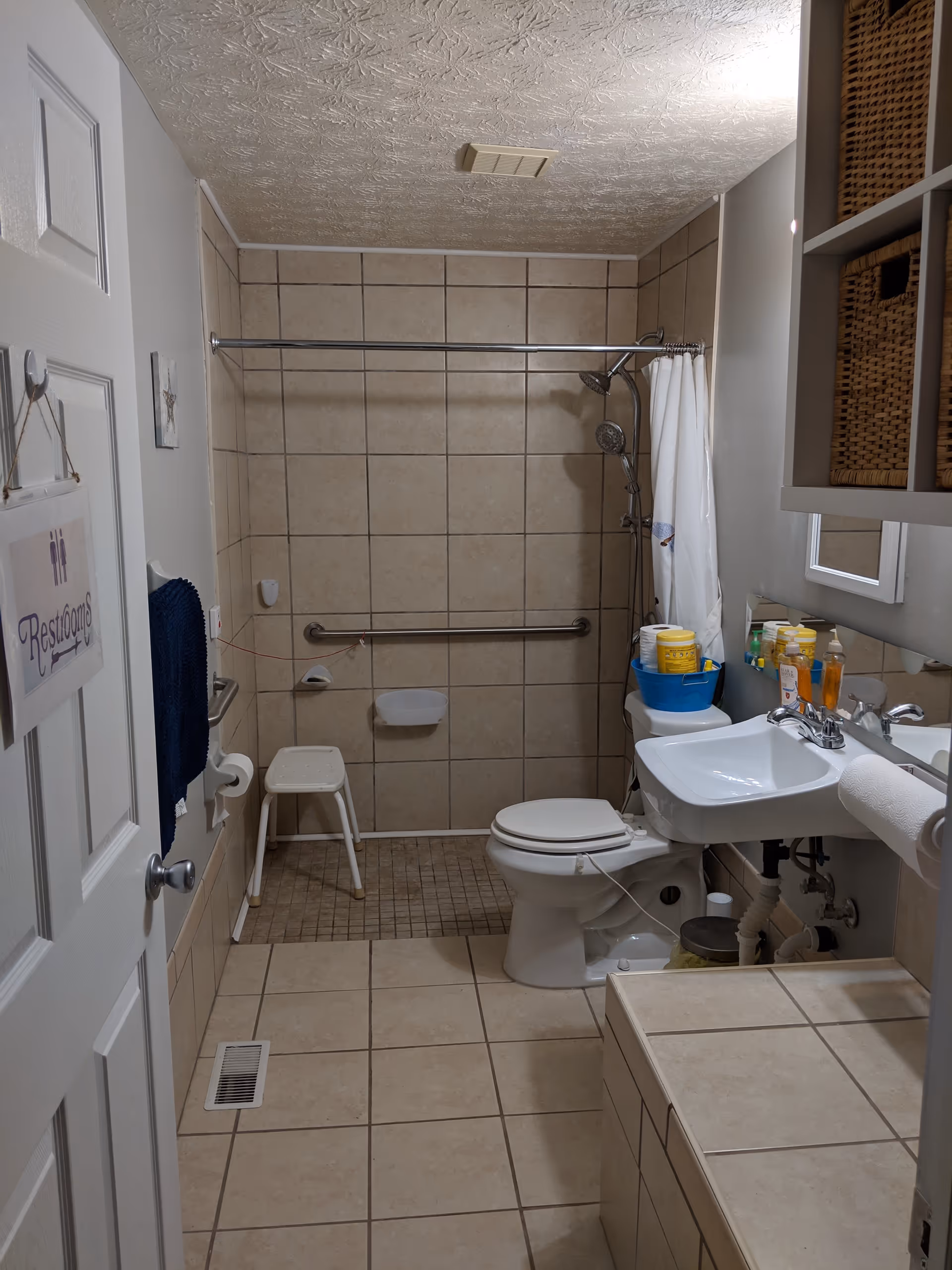 A bathroom with beige tiled walls and floor. There is a white toilet with a blue basket on top holding cleaning supplies, a white sink with a mirror above it, and a shower area with a white shower curtain and a small white stool. A grab bar is installed on the shower wall. A towel hangs on the wall near the door, which has a sign that reads 'Restrooms'.