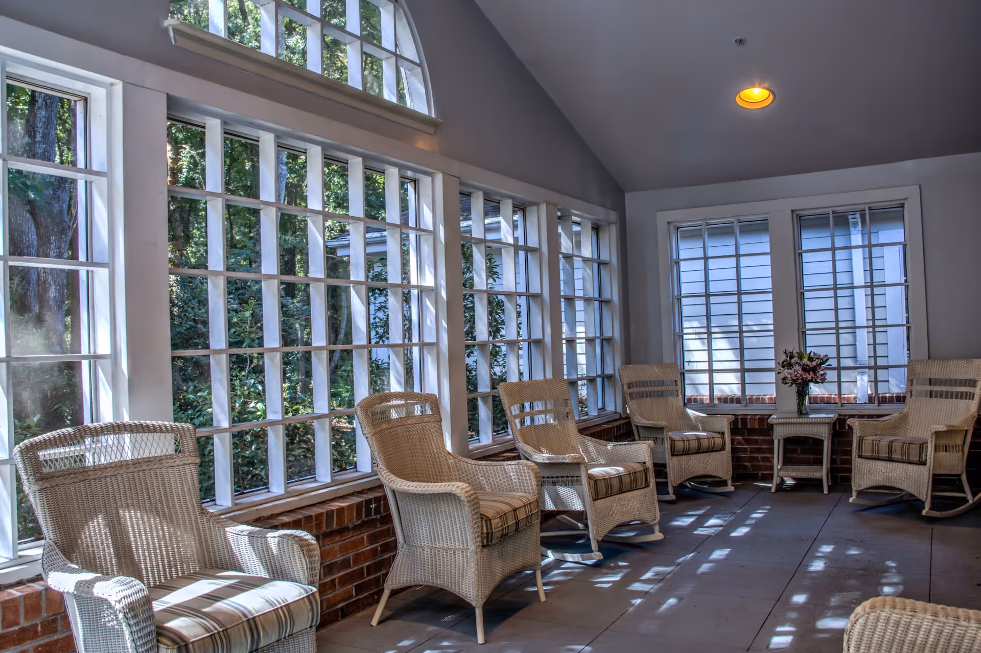 A sunlit enclosed porch area with large windows and white wicker chairs arranged along the walls. There is a small table with a vase of flowers in the corner. The floor is tiled and the walls are painted light gray.