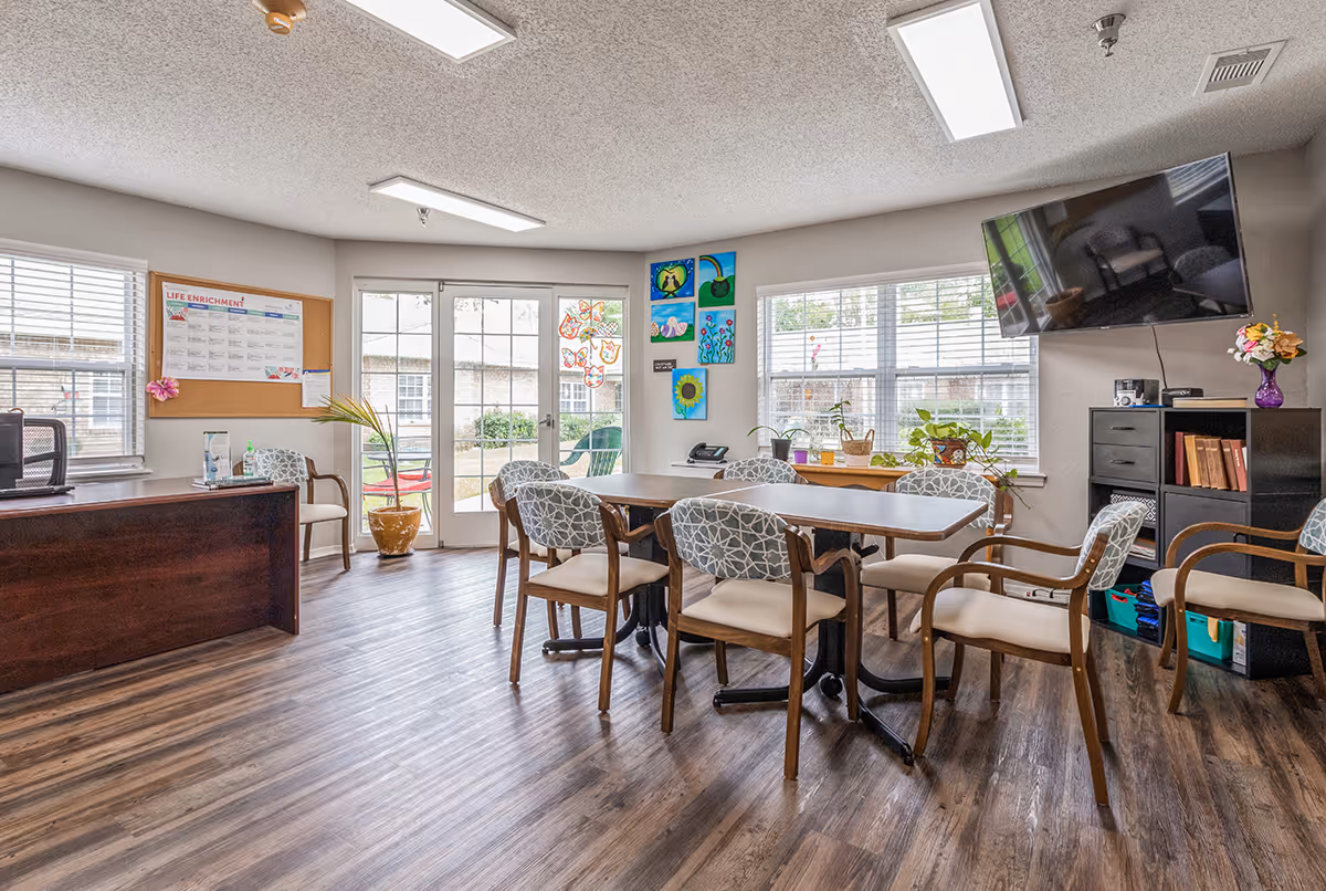 Bright communal dining/activity room with a large table and chairs, windows, wall art, and a mounted TV.
