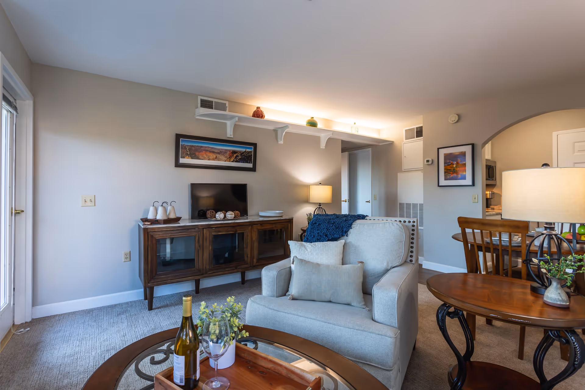 A cozy living room in a senior living facility featuring a comfortable armchair with cushions, a wooden coffee table with a bottle of wine, two wine glasses, and a small plant. A wooden TV stand with a flat-screen TV and decorative items is against the wall. The room has soft lighting from a table lamp and natural light coming through a glass door. A dining area with wooden chairs and a table is visible through an archway.