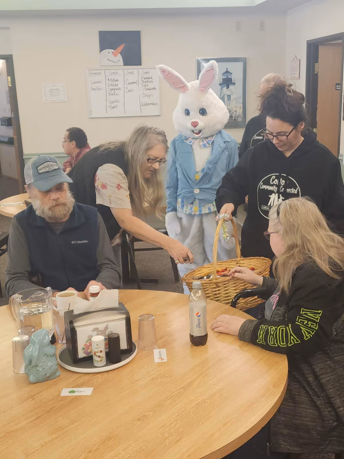 People seated around a round table in a communal dining area as someone in an Easter bunny costume offers treats from a basket.