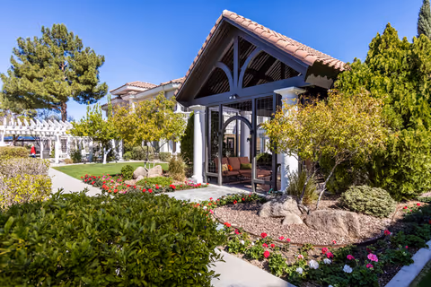 Outdoor view of a senior living facility with a covered seating area featuring cushioned chairs, surrounded by well-maintained landscaping including bushes, flowering plants, and trees under a clear blue sky.