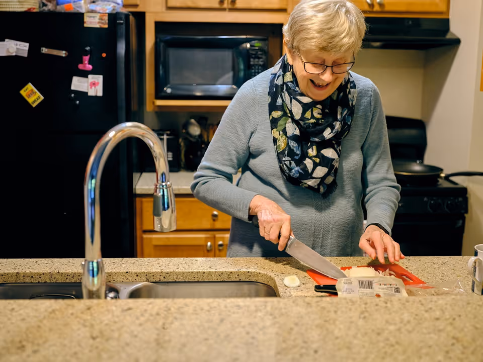 An elderly woman with short blonde hair and glasses is smiling while chopping an onion on a red cutting board in a kitchen. She is wearing a light blue sweater and a patterned scarf. The kitchen has a granite countertop, a stainless steel sink with a curved faucet, wooden cabinets, a black refrigerator with magnets, a microwave, and a stove with a frying pan.