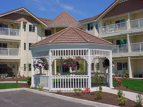 A white gazebo with hanging flower baskets sits in the courtyard in front of a multi-story retirement community building.