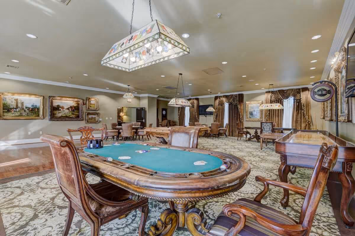 Ornate senior living game room with a poker table in the foreground, shuffleboard and multiple seating areas under decorative pendant lights.