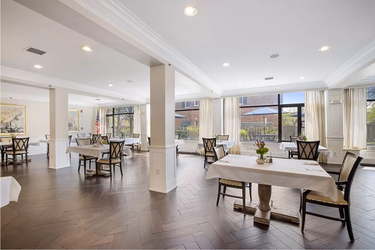 A bright and spacious dining room with several tables covered in white tablecloths, each set with chairs and small flower arrangements. Large windows allow natural light to fill the room, and there are white curtains and a wooden floor with a herringbone pattern.