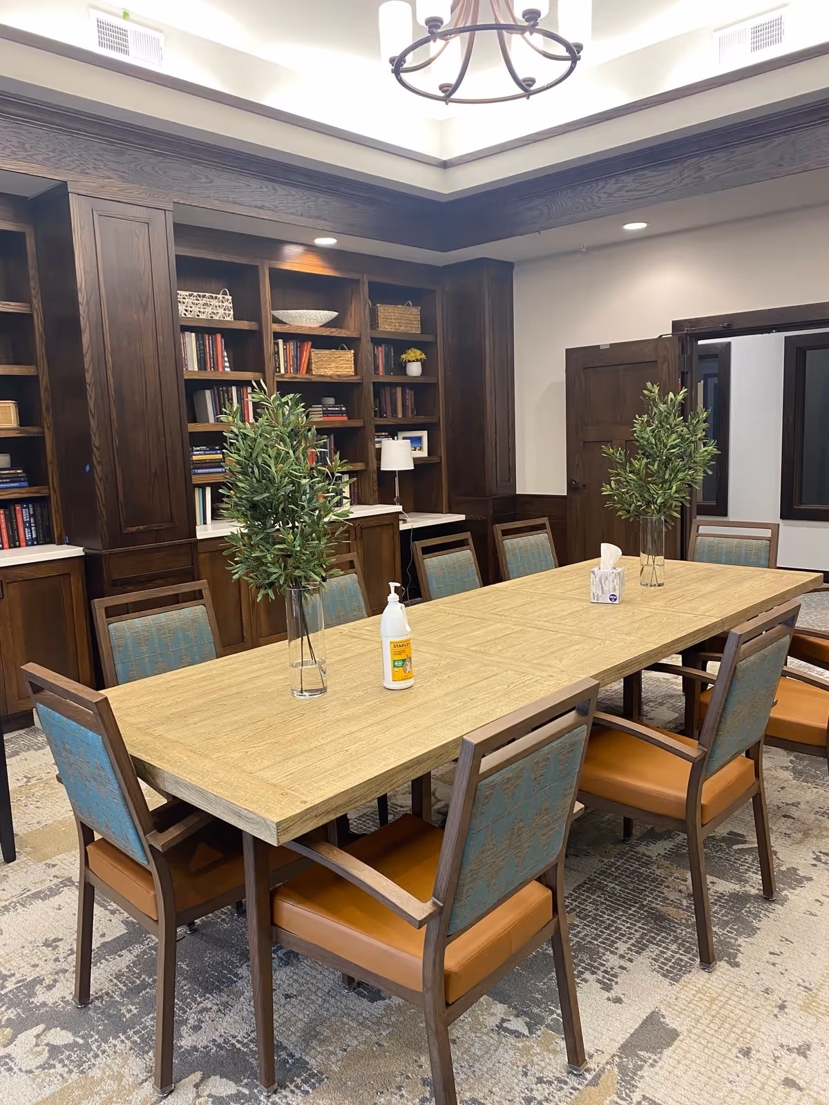 A well-lit meeting or dining room with a long wooden table surrounded by eight chairs with blue patterned backs and brown seats. The table has two glass vases with green leafy plants, a bottle of hand sanitizer, and a box of tissues. The room features dark wood built-in bookshelves filled with books and decorative items, a beige patterned carpet, and a chandelier hanging from the ceiling.