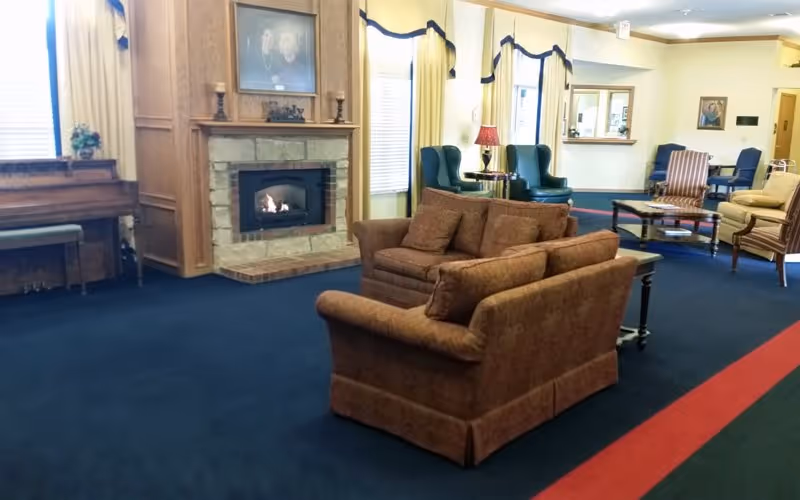 A cozy living room area with a stone fireplace, two brown upholstered sofas facing each other, a piano on the left side, and several armchairs and tables arranged around the room. The room has blue carpet with a red stripe and yellow curtains on the windows.