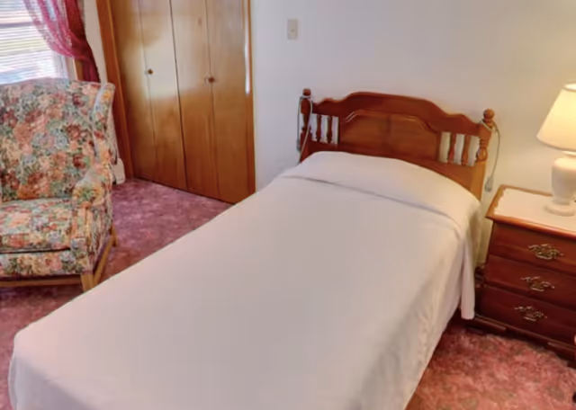A bedroom with a single wooden bed covered with a white sheet, a wooden nightstand with a lamp on it, a floral upholstered armchair, a window with red curtains, and a wooden closet with double doors.