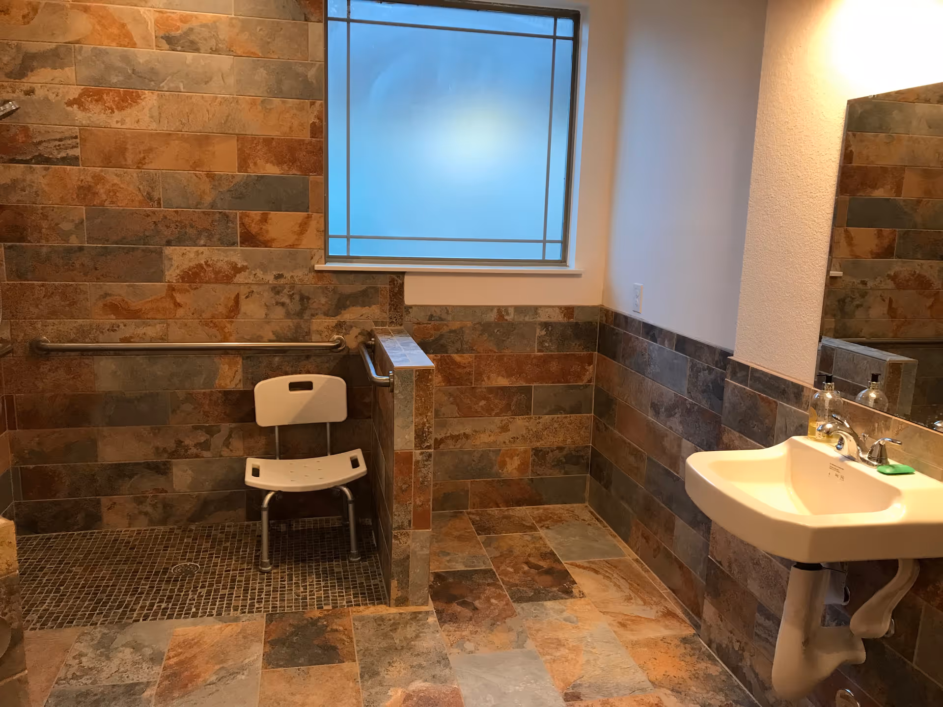 A bathroom with multi-colored stone tiles on the floor and walls, featuring a walk-in shower area with a white shower chair and grab bars, a frosted window, and a white wall-mounted sink with a mirror above it.