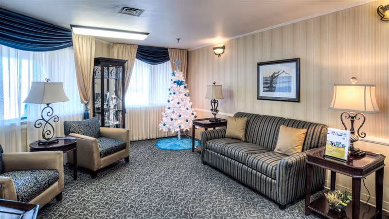 A cozy living room area with a striped sofa, two armchairs, two side tables with lamps, a white decorated Christmas tree, a glass display cabinet, and framed artwork on the wall. The room has patterned carpet and windows with curtains and valances.