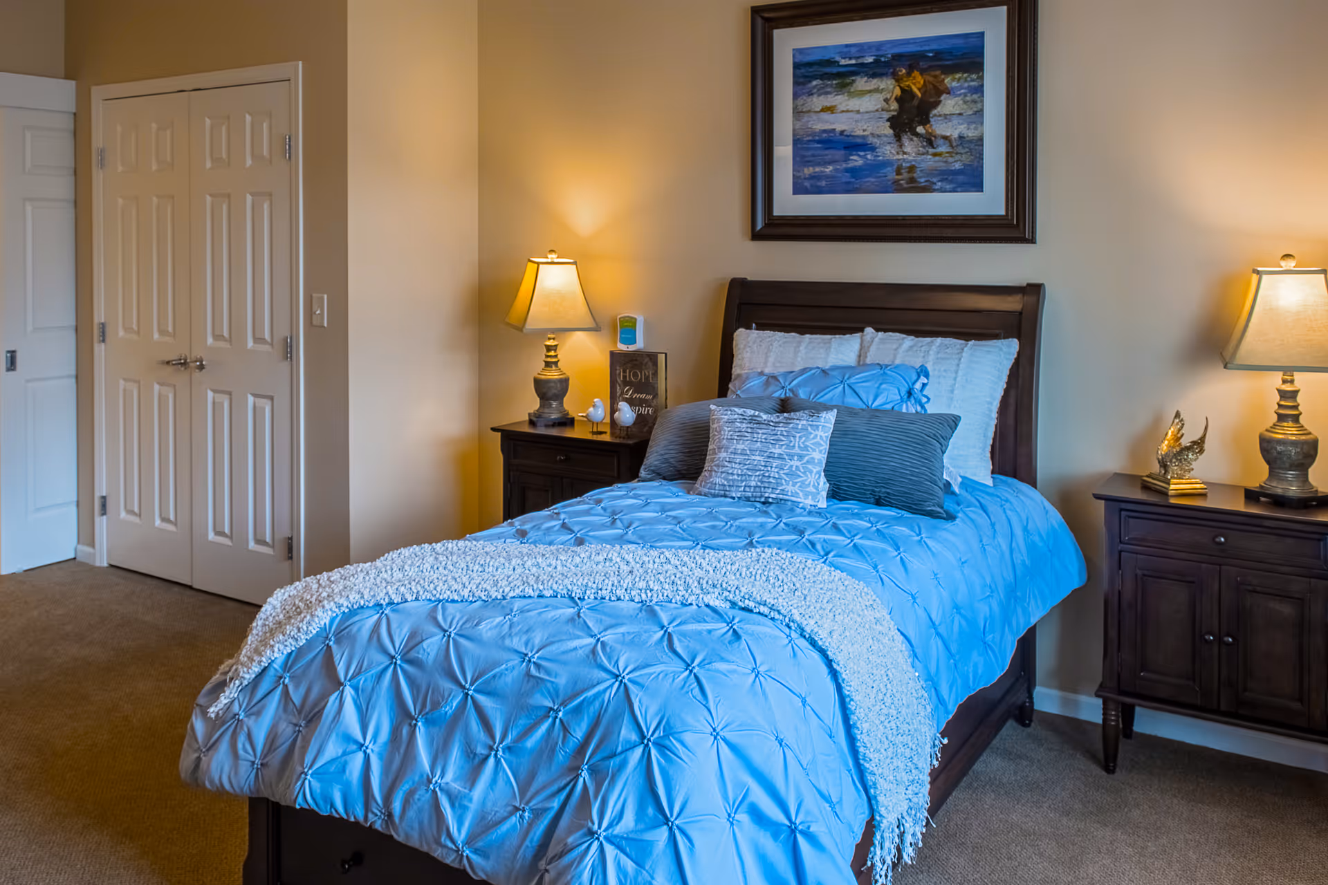 A cozy bedroom with a single bed dressed in a light blue comforter and several pillows. There are two dark wooden nightstands on either side of the bed, each with a lamp. A framed painting hangs on the wall above the bed. The room has beige walls and carpeted flooring, with a closed double-door closet visible on the left side.