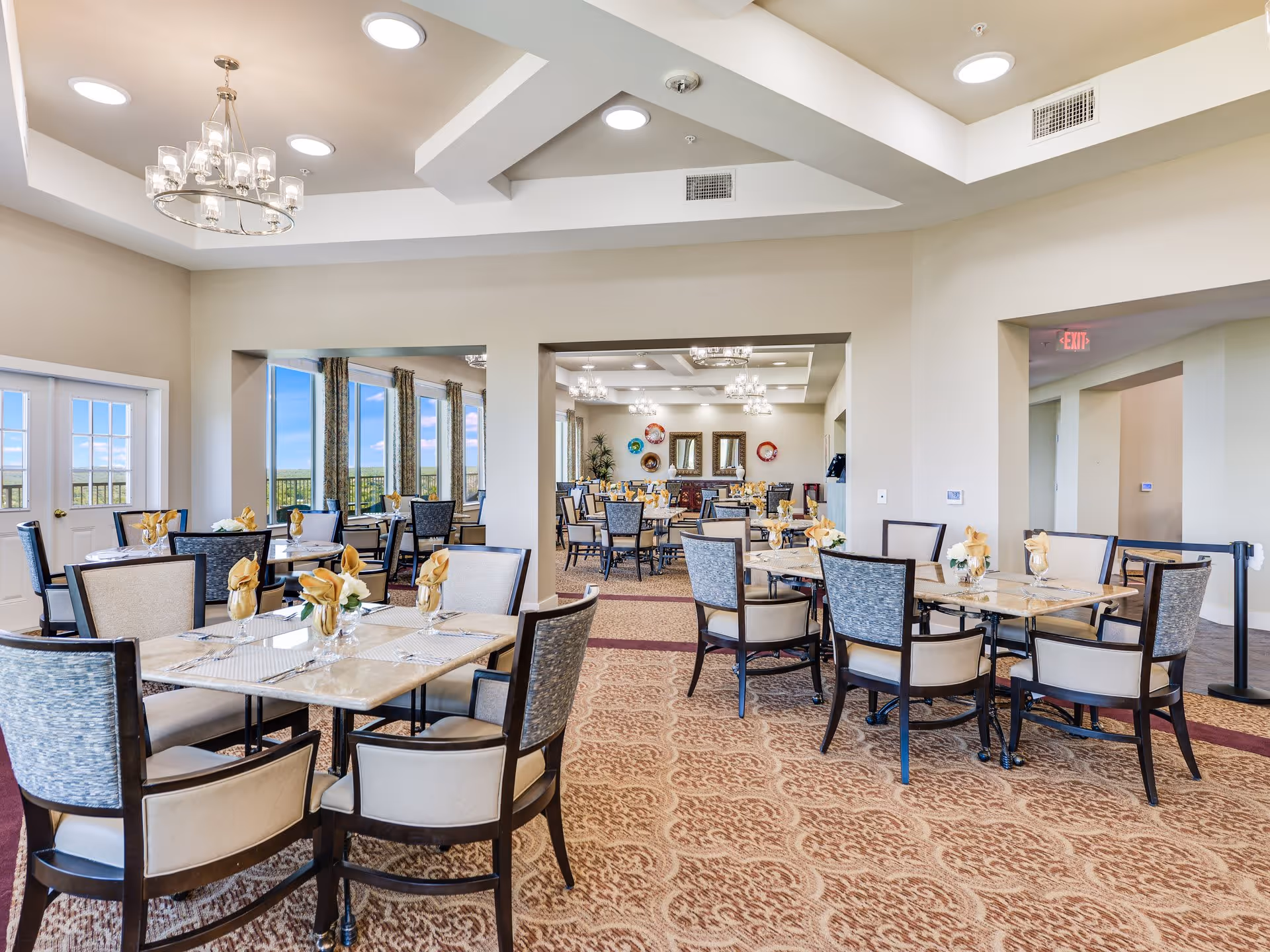 A spacious and well-lit dining room in a senior living facility with multiple tables set with beige napkins and glassware. The room features large windows with a scenic view, chandeliers hanging from a tray ceiling, and patterned carpet flooring.