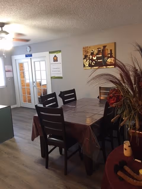 Interior view of a dining area with a rectangular table covered with a plastic tablecloth and surrounded by six dark wooden chairs. On the right side, there is a decorative arrangement with tall dried plants and a candle on a small round table. The background shows a wall with a framed artwork of vases and a set of double glass doors with white frames. The floor is wood laminate, and a ceiling fan with lights is visible.