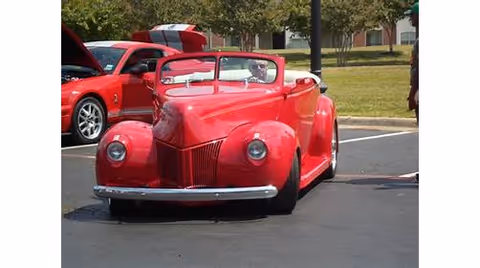 A bright red vintage convertible car parked on an asphalt surface with a person sitting inside. Another red car with its hood open is visible in the background along with trees and a grassy area.