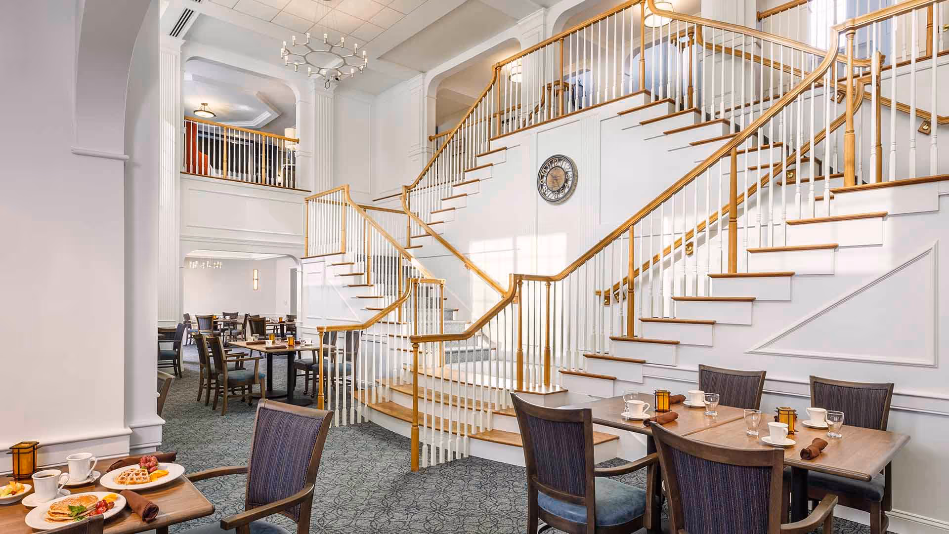 A spacious dining area in a senior living facility featuring multiple tables set with cups, plates, and napkins. The room has a large, elegant staircase with wooden handrails and white balusters leading to an upper level. The walls are white with decorative molding, and the carpet is patterned in a muted color. The space is well-lit with natural light and ceiling fixtures.