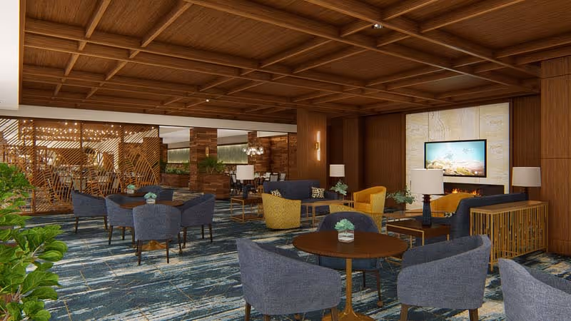 A spacious and elegantly designed common area with multiple seating arrangements including blue and yellow armchairs around small round tables. The room features a wooden coffered ceiling, a patterned blue carpet, and a fireplace with a mounted flat-screen TV above it. There are decorative plants and modern lamps placed on tables, and a wooden partition with a geometric design separates part of the space.