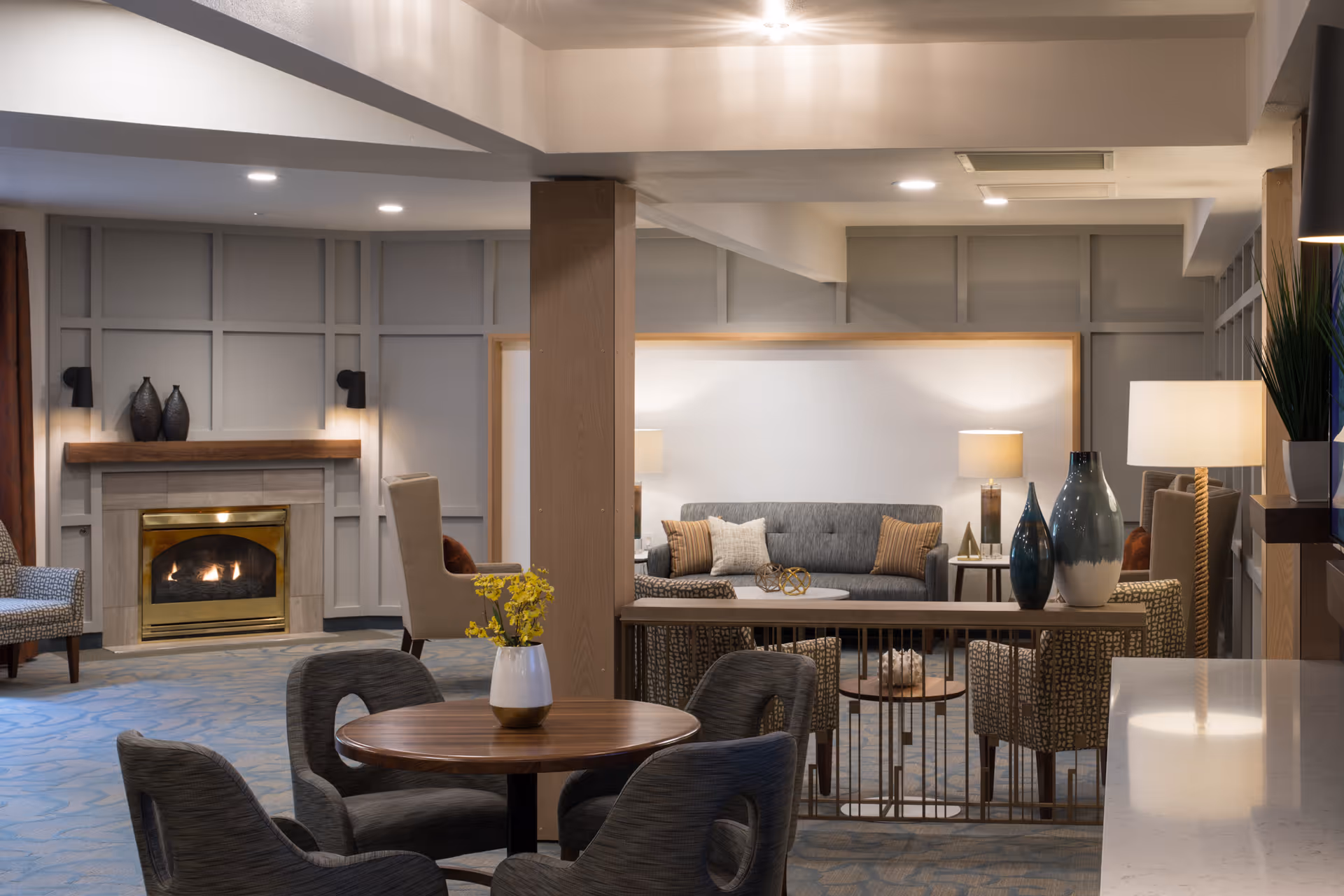 A cozy communal living room with a lit fireplace, sofas, chairs, lamps, and decorative vases.