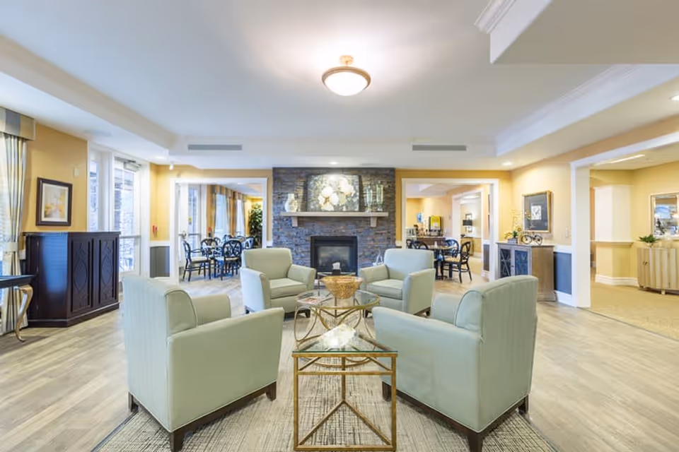 A bright and spacious living room area in a senior living facility with four light green armchairs arranged around a glass coffee table on a rug. The room features a stone fireplace with decorative items on the mantel, light yellow walls, wood flooring, and multiple doorways leading to other rooms with dining tables and chairs visible.