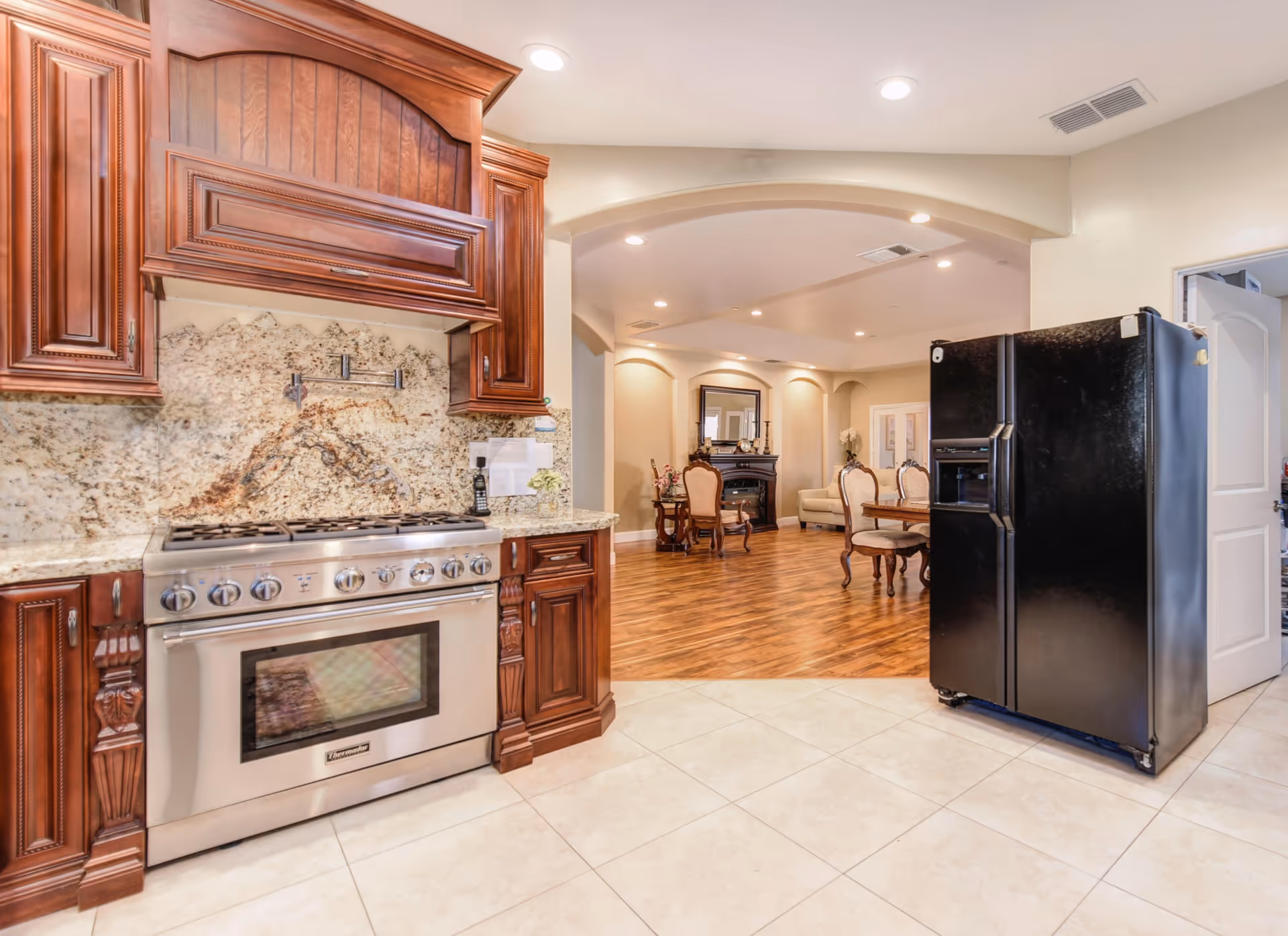 Spacious kitchen with dark wooden cabinets, a stainless steel range, and a black refrigerator opening to a dining/living area through an arched doorway.