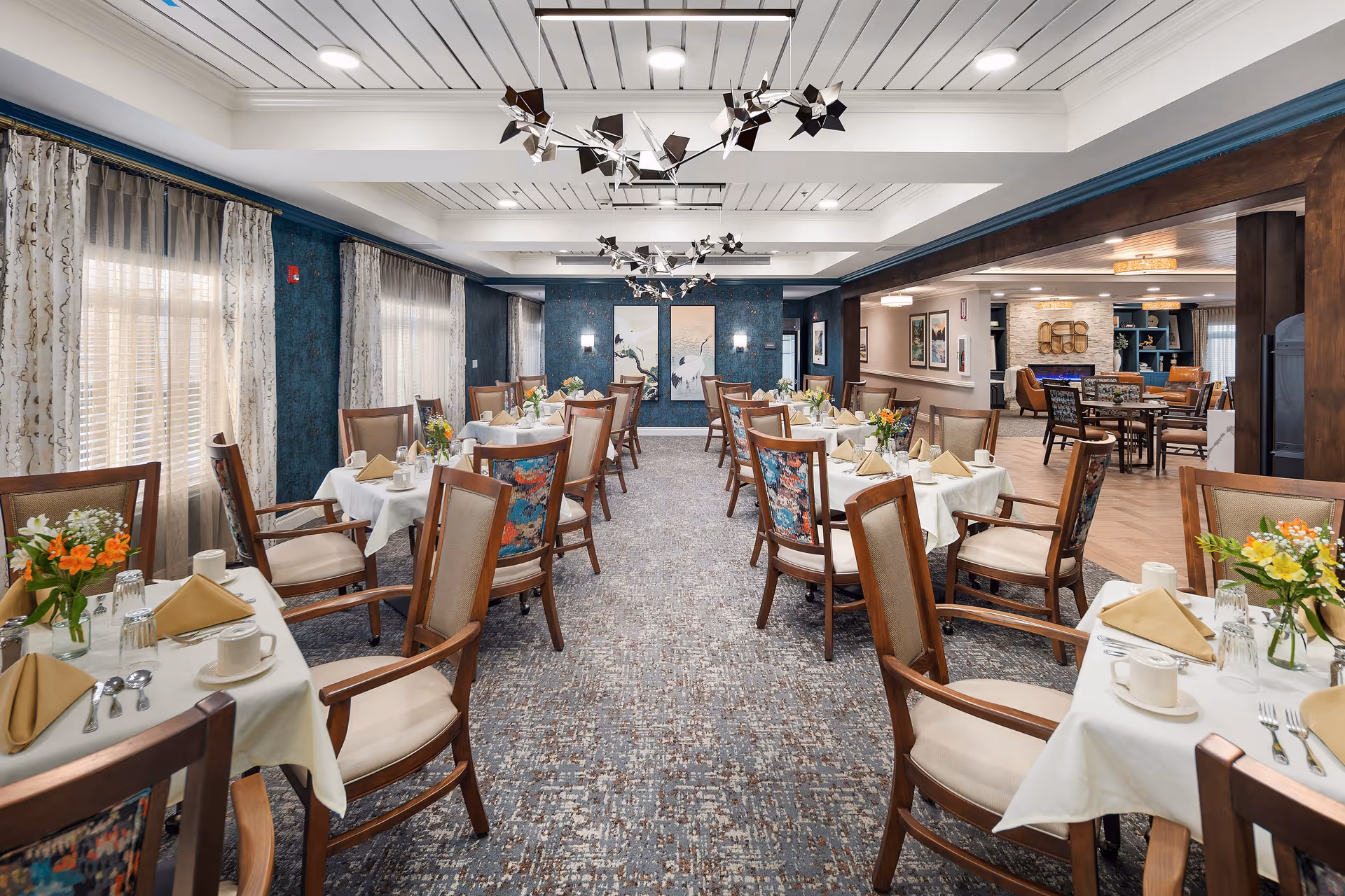 A bright and elegant dining room with multiple tables set with white tablecloths, beige napkins, cups, and silverware. Each table has a small floral centerpiece. The room features large windows with sheer curtains, modern ceiling lights, and decorative wall art at the far end. Adjacent to the dining area is a cozy lounge space with armchairs and a fireplace.