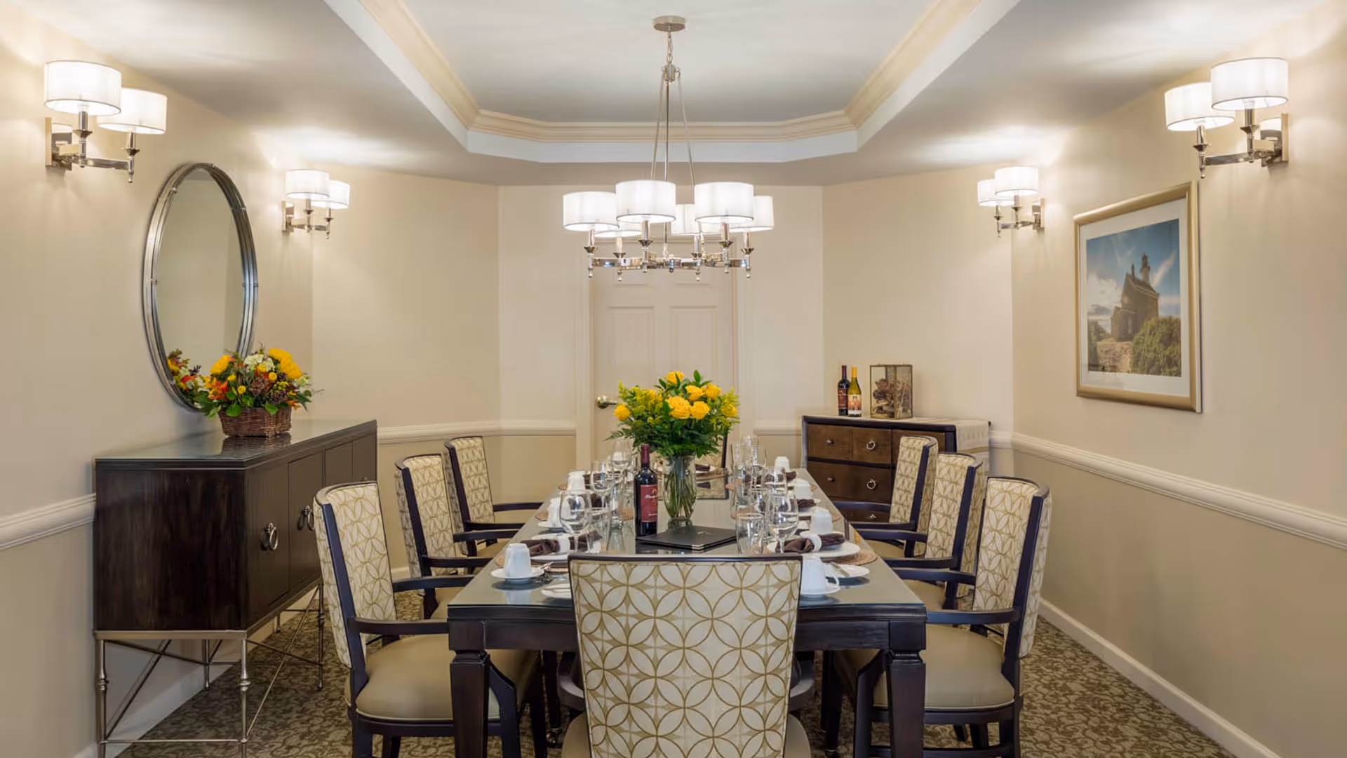 A formal dining room with a long rectangular table set for a meal. The table has ten upholstered chairs with a circular pattern, a vase of yellow flowers in the center, wine bottles, glasses, and place settings. The room has beige walls, a patterned carpet, wall sconces with white lampshades, a round mirror above a dark wooden sideboard with a flower arrangement, and a framed picture on the opposite wall.