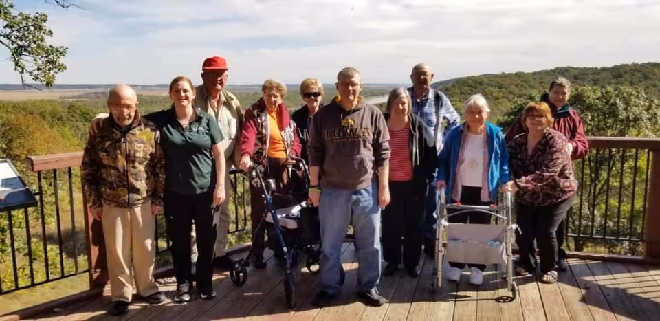 A group of elderly people and a caregiver standing together on a wooden deck with a scenic view of trees and hills in the background. Some individuals are using walkers for support.