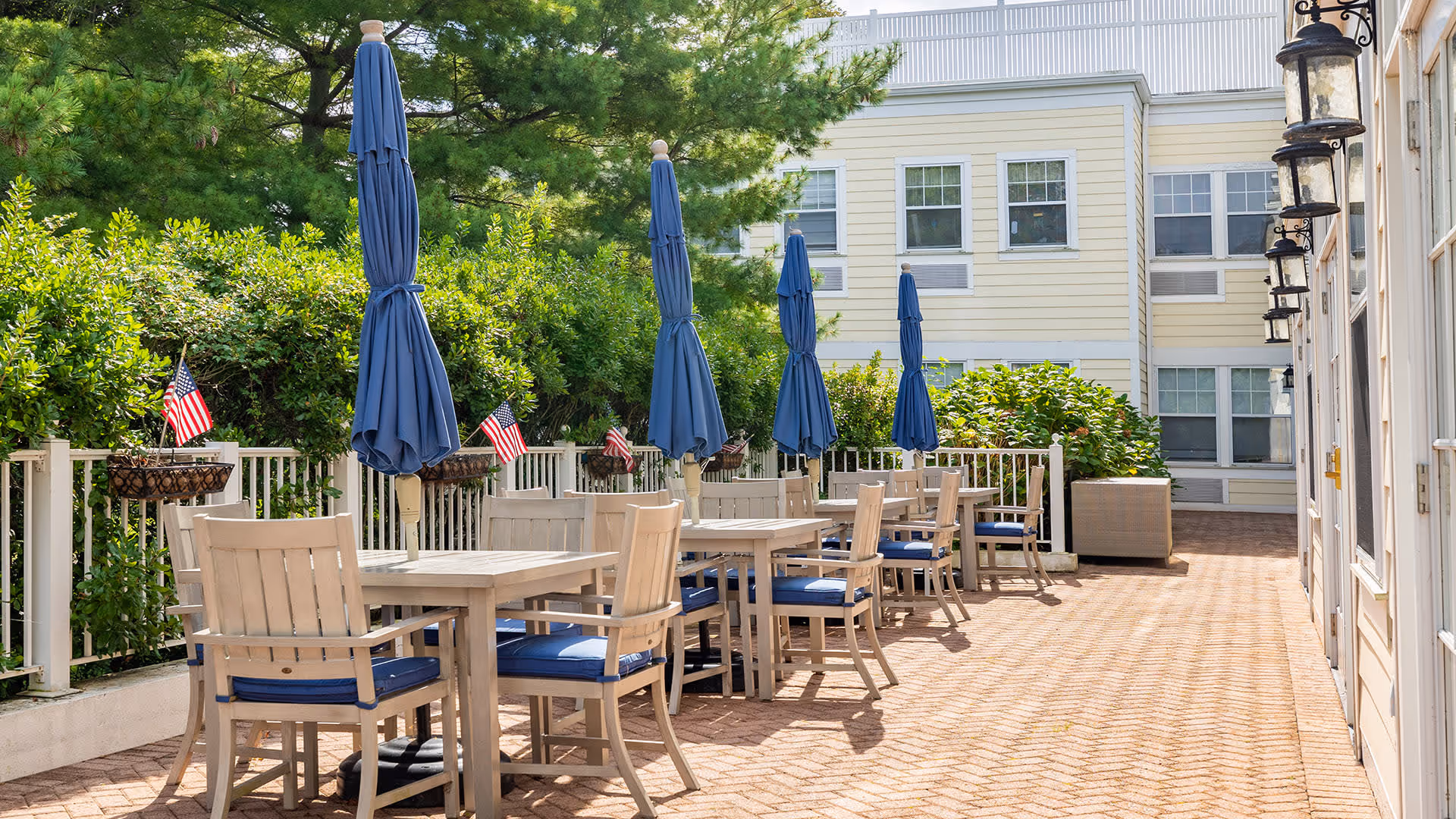 Outdoor patio area with several wooden tables and chairs featuring blue cushions and closed blue umbrellas. The patio is surrounded by greenery and small American flags are placed in flower baskets along the white railing. A light yellow building with multiple windows is visible in the background.