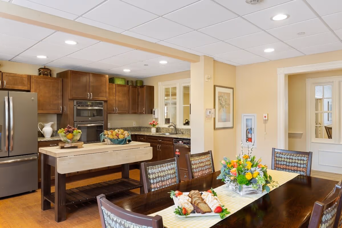 Bright communal kitchen and dining area with a wooden island, stainless steel appliances, and a dining table set with flowers and food.