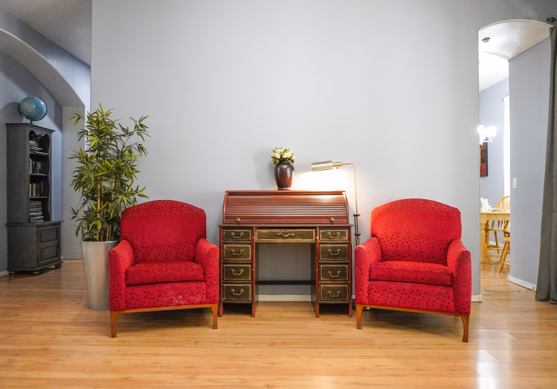 Two red upholstered armchairs flank a vintage roll-top desk with a lamp and potted plant in a bright living room with hardwood floors.
