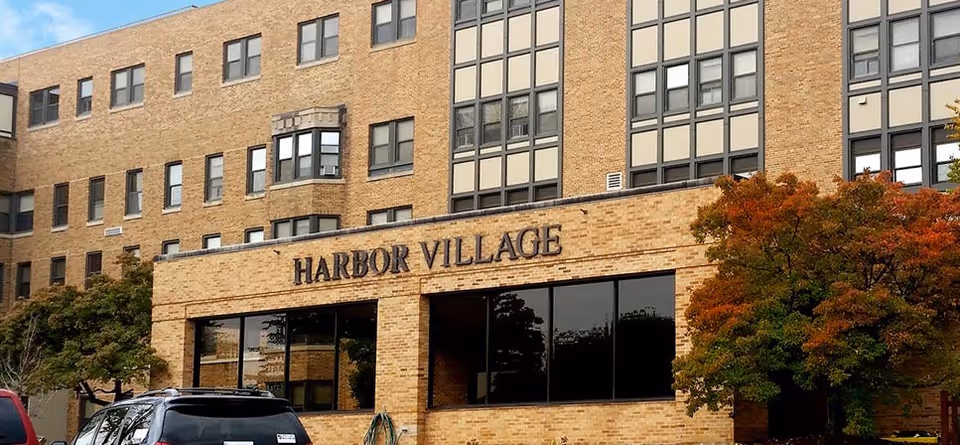 Exterior view of a multi-story brick building with large windows and a sign that reads 'HARBOR VILLAGE'. There are trees with autumn foliage and a parked car visible in front of the building.