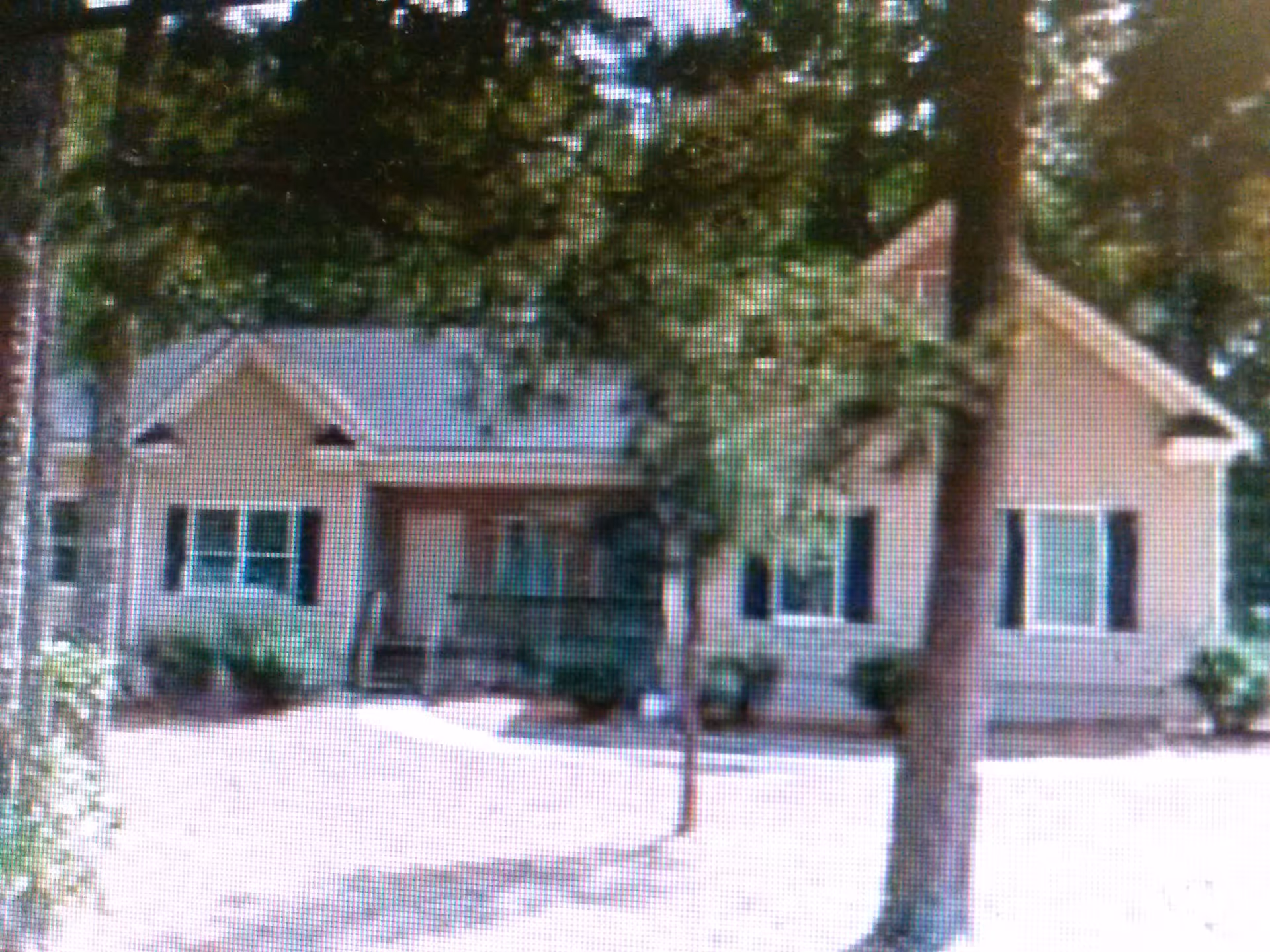 Exterior view of a single-story house with a front porch, surrounded by trees and shrubs, under daylight.