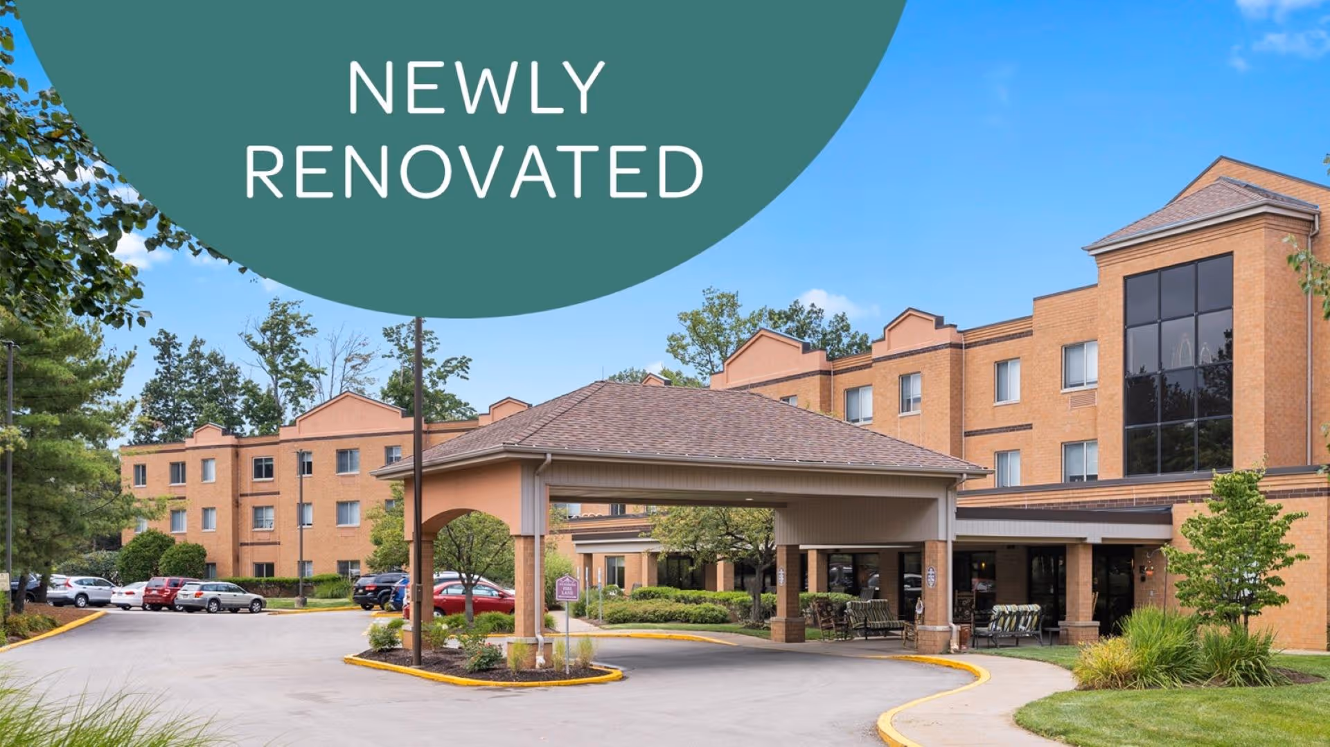 Exterior view of a newly renovated senior living facility with a covered entrance, multiple parked cars, and a three-story brick building surrounded by trees and landscaping under a clear blue sky.