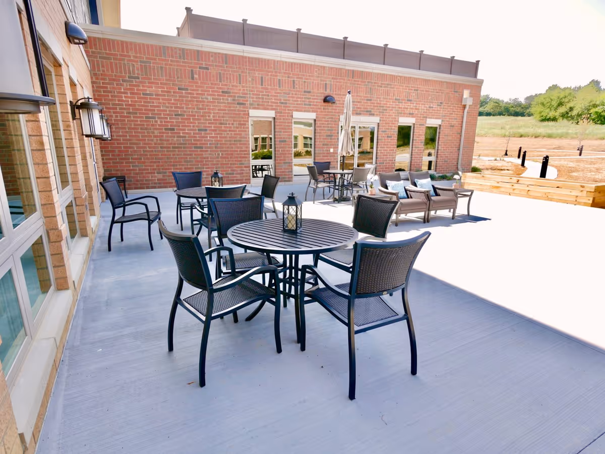 Outdoor patio area with several black metal tables and chairs arranged on a concrete surface next to a brick building. There are lanterns on the tables, a closed umbrella, and a seating area with cushioned chairs and small tables. The background shows a grassy field and trees.