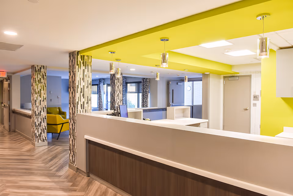 Interior view of a nursing and rehabilitation center reception area with a modern design. The space features a long reception desk with a computer, pendant lights hanging from a bright yellow ceiling section, patterned columns, and wood-patterned flooring. There is a hallway leading to other rooms and a yellow armchair visible in the background.