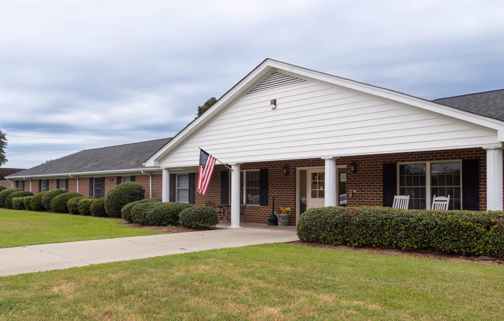Exterior view of a single-story brick building with white columns and a gabled roof. The building has several windows with black shutters, an American flag hanging near the entrance, and a concrete walkway leading to the door. There are neatly trimmed bushes and a well-maintained lawn in front of the building under a cloudy sky.