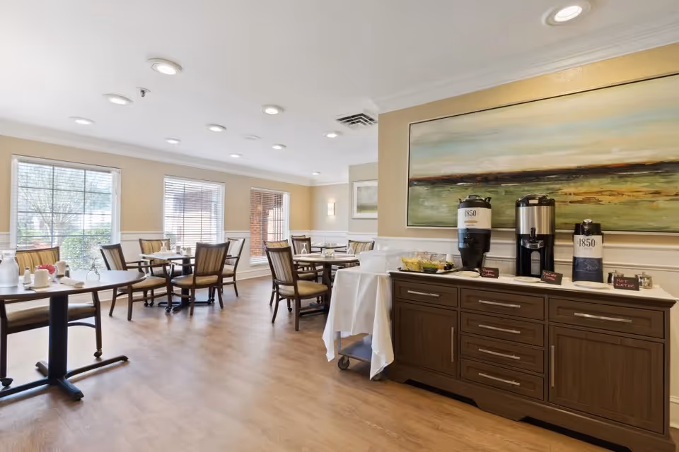 A bright dining room with several tables and chairs arranged neatly. Large windows allow natural light to fill the space. On the right side, there is a wooden sideboard with coffee dispensers labeled '1850' and other beverage containers. Above the sideboard hangs a large landscape painting.