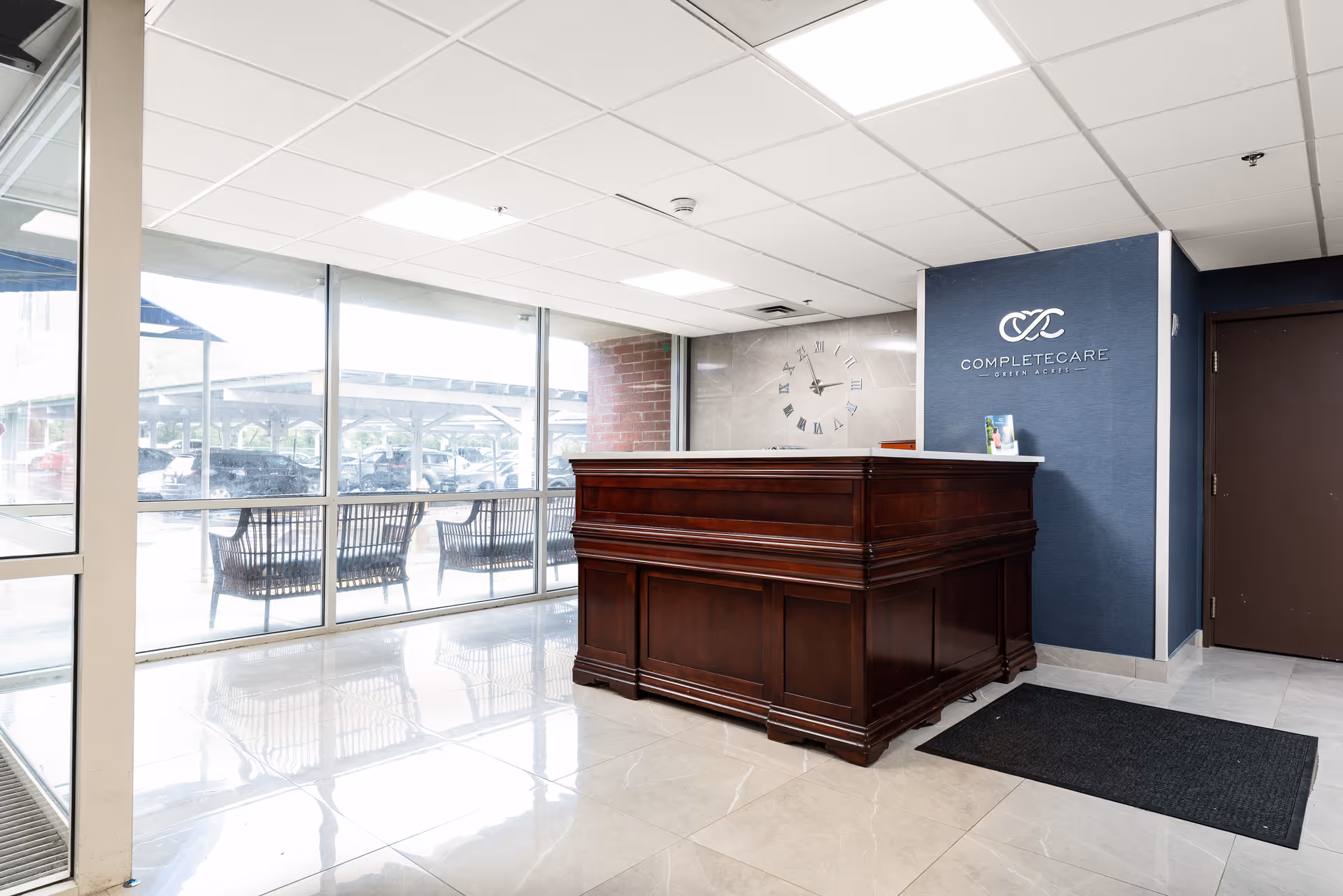 Reception area of Complete Care at Green Acres featuring a large wooden front desk, a wall clock with Roman numerals, a blue accent wall with the facility's logo, and large windows showing outdoor seating and a parking area.