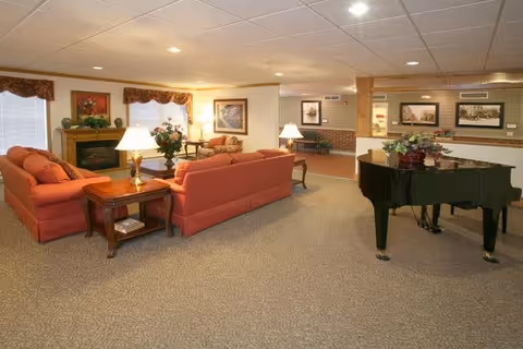 A spacious living room area with two orange sofas arranged around a wooden coffee table and side tables with lamps. There is a fireplace with a painting above it, windows with valances, and a black grand piano with a flower arrangement on top. The walls have framed pictures and the floor is carpeted.